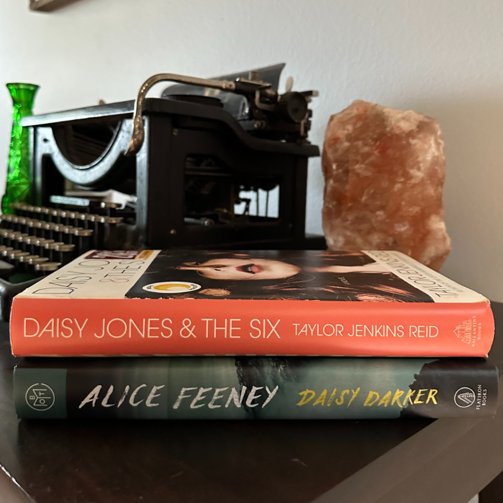Two books, Daisy Jones & the Six by Taylor Jenkins Reid and Daisy Darker by Alice Feeney, stacked on a table with a vintage typewriter, salt lamp, and green vases
